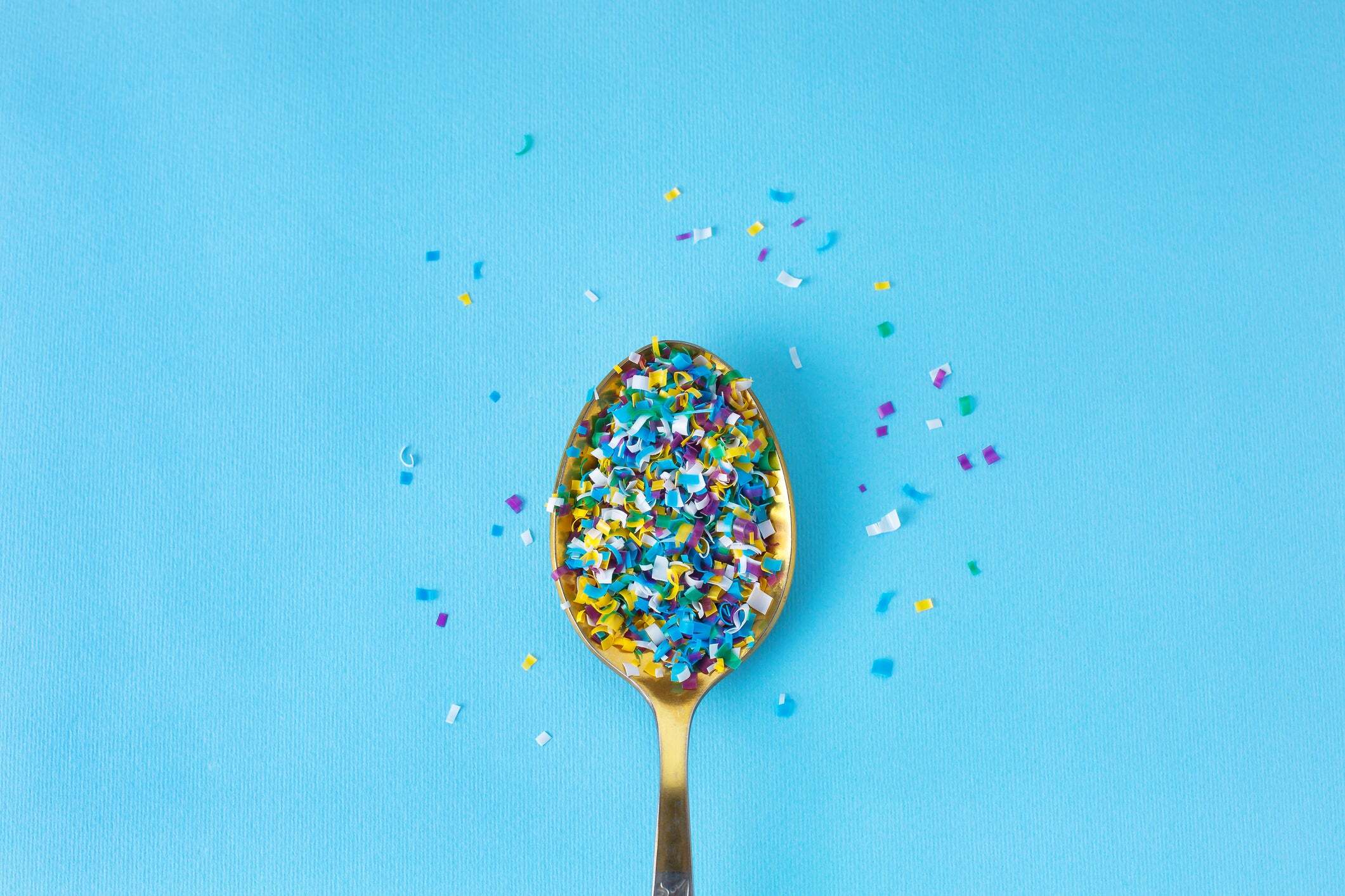 Spoon filled with colorful microplastic fragments on a blue background, illustrating plastic particles commonly found in everyday materials.