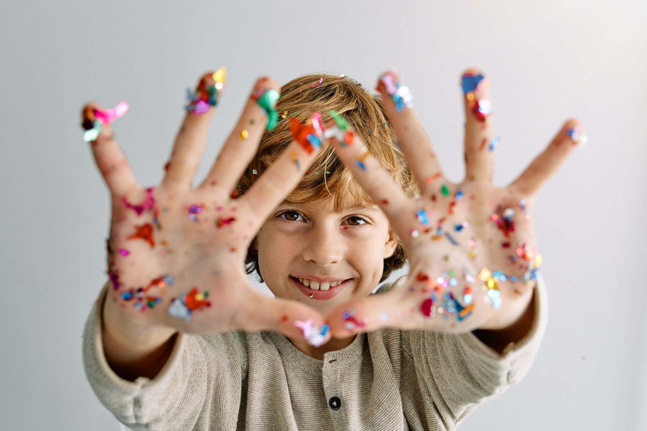 Child smiling with hands covered in colorful craft materials, showing the hands-on nature of classroom projects using school adhesives.